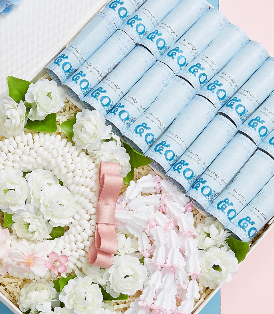 An elegantly decorated box from Basketeer, adorned with luxurious detailing, features carefully rolled blue parchment documents arranged methodically on one side. The other side showcases an opulent floral arrangement of pristine white blooms, lush green foliage, and delicate pink accents. Completing this exquisite presentation is a sophisticated pink ribbon, subtly bearing the Basketeer logo for an added touch of refinement and brand prestige.