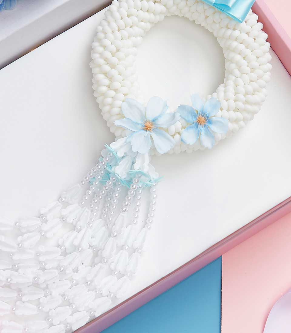 A luxurious decorative white flower garland embellished with delicate light blue flowers and cascading strands of pristine white and azure beads is elegantly laid on a pastel-colored surface, showcasing serene hues of pink, blue, and soft white. This intricately crafted garland is crowned with an exquisite blue ribbon bearing the distinguished Basketeer logo, evoking a sense of refined elegance and inviting beauty.