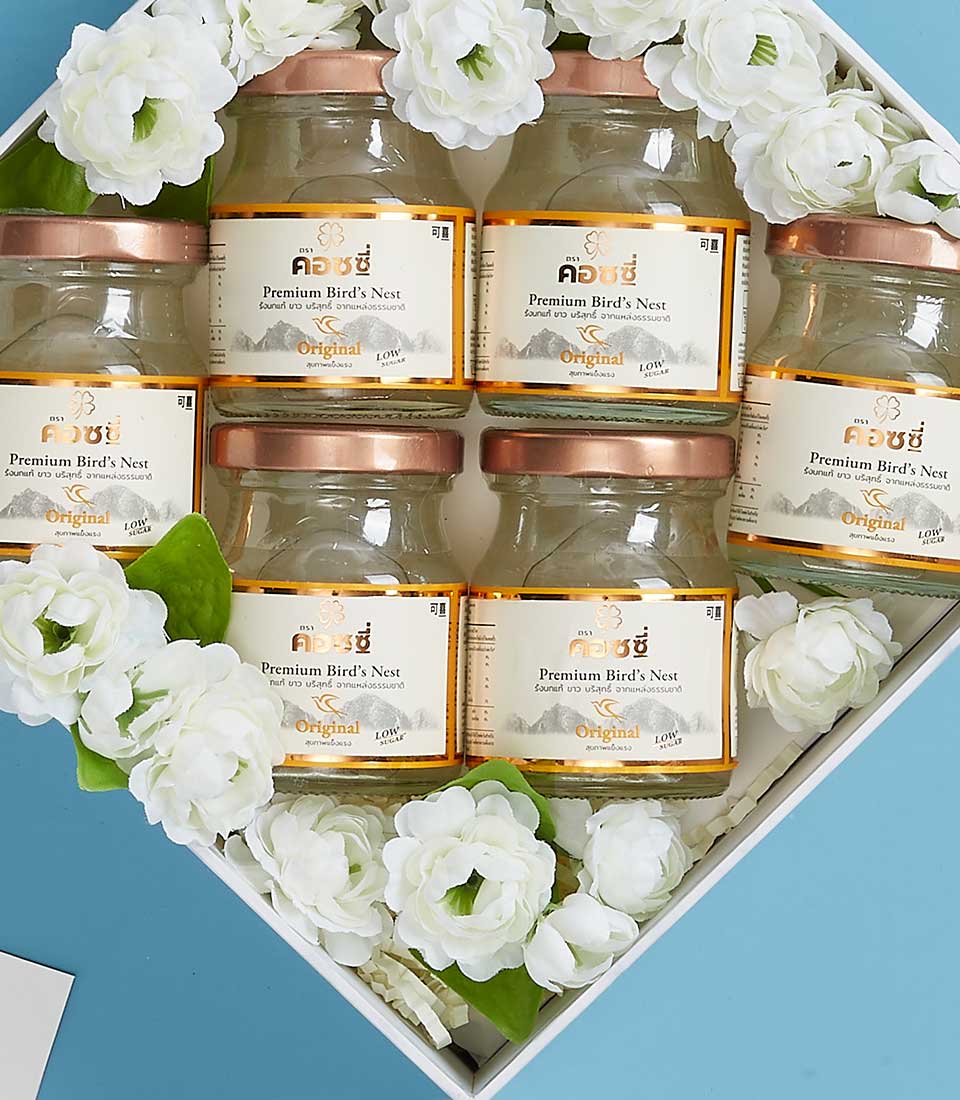 A sumptuous gift set from Basketeer, featuring six jars of premium bird's nest with pristine white labels and sophisticated brown lids, beautifully displayed in an exquisite white box. Each jar nestles amidst delicate white artificial flowers, radiating elegance and luxury. The arrangement is accentuated by a light blue backdrop, enhancing the inviting allure of the scene. A ribbon bearing the prestigious Basketeer logo adds a final touch of refinement.