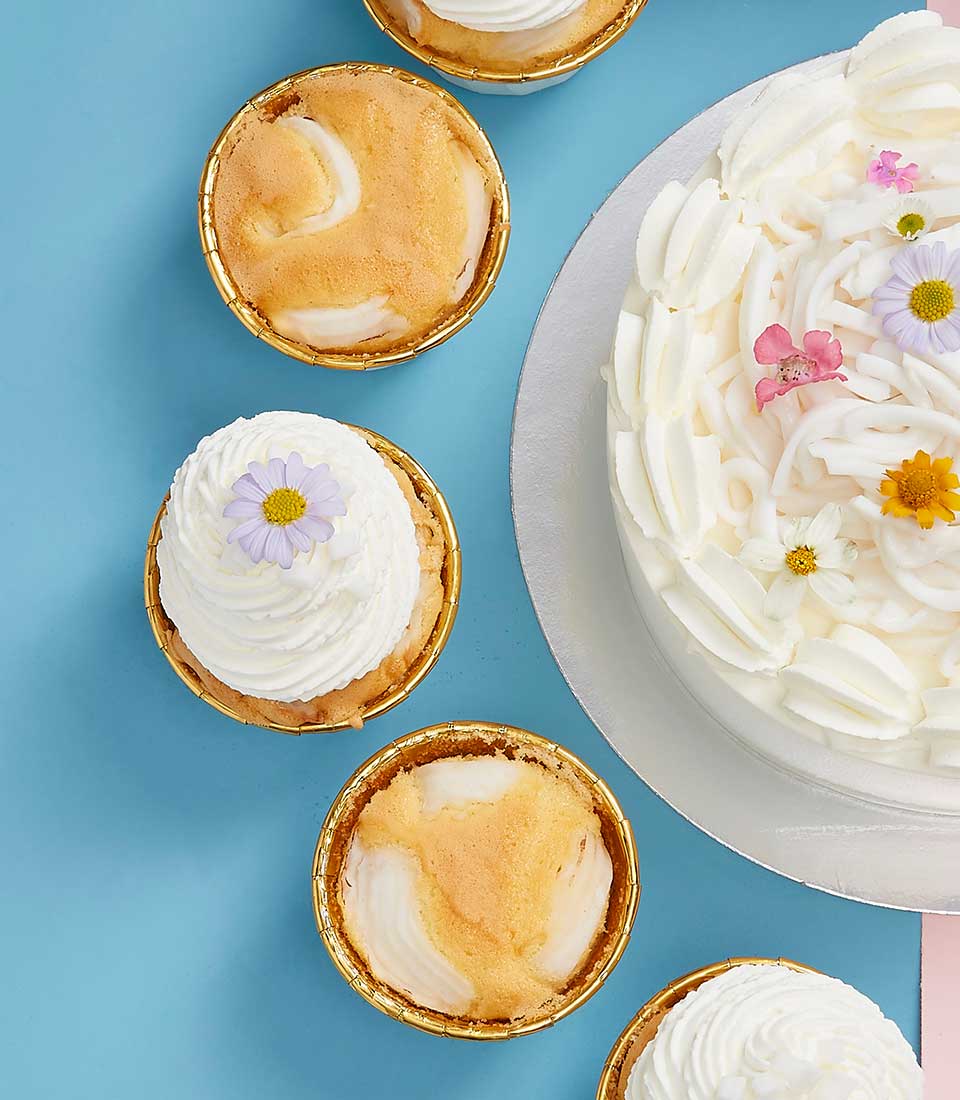 A decadent white-frosted coconut cake, elegantly adorned with an array of vibrant flowers, takes center stage on a gleaming silver platter. Encircling this masterpiece are four exquisite tarts; two mirror the main cake's floral embellishments and swirled white frosting, while the other two boast a luxurious golden-brown top. These delightful treats rest on a soft blue surface, creating a visually stunning display that epitomizes Basketeer’s commitment to luxury and indulgence. The presence of a ribbon or box bearing the distinguished Basketeer logo further enhances the scene with an aura of sophistication and exclusivity.