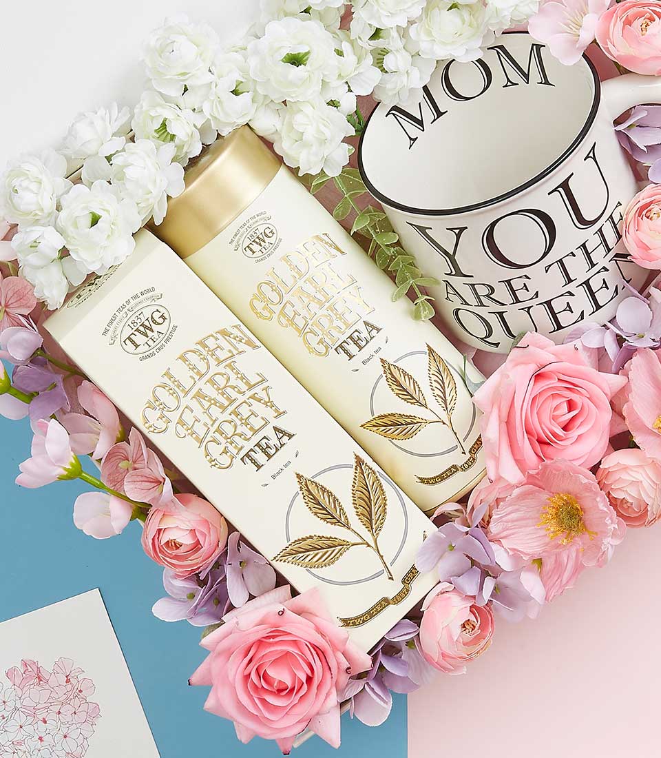 Two elegantly cylindrical containers of TWG Golden Earl Grey Tea are nestled amidst a lavish arrangement of pink and white flowers, exuding luxury and refinement. To the right, a pristine white cup with the heartfelt message 