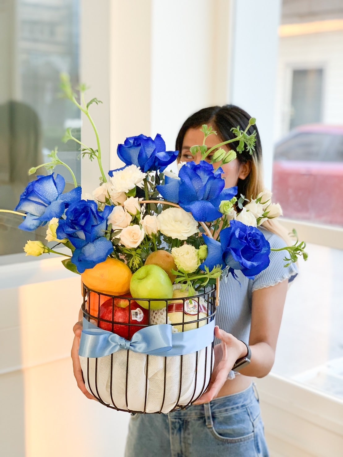 A person holds a white wire fruit basket filled with colorful apples, oranges, and pears, alongside a vibrant arrangement of bright flowers. The basket is decorated with a light blue ribbon and is set against a softly lit indoor background.
