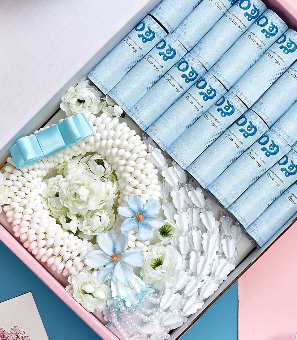A meticulously curated Basketeer gift box exudes luxury and charm, featuring plush blue rolled-up towels adorned with a delicate light blue satin bow. Pristine white floral garlands intertwine with ethereal white flowers accented by soft blue petals. The interior of the box is elegantly lined with a gentle pink border, enhancing the overall aesthetic. A hint of pastel pink and blue paper peeks out, adding a touch of sophisticated whimsy. Basketeer's logo on a ribbon or box subtly emphasizes the brand's commitment to opulence and elegance.