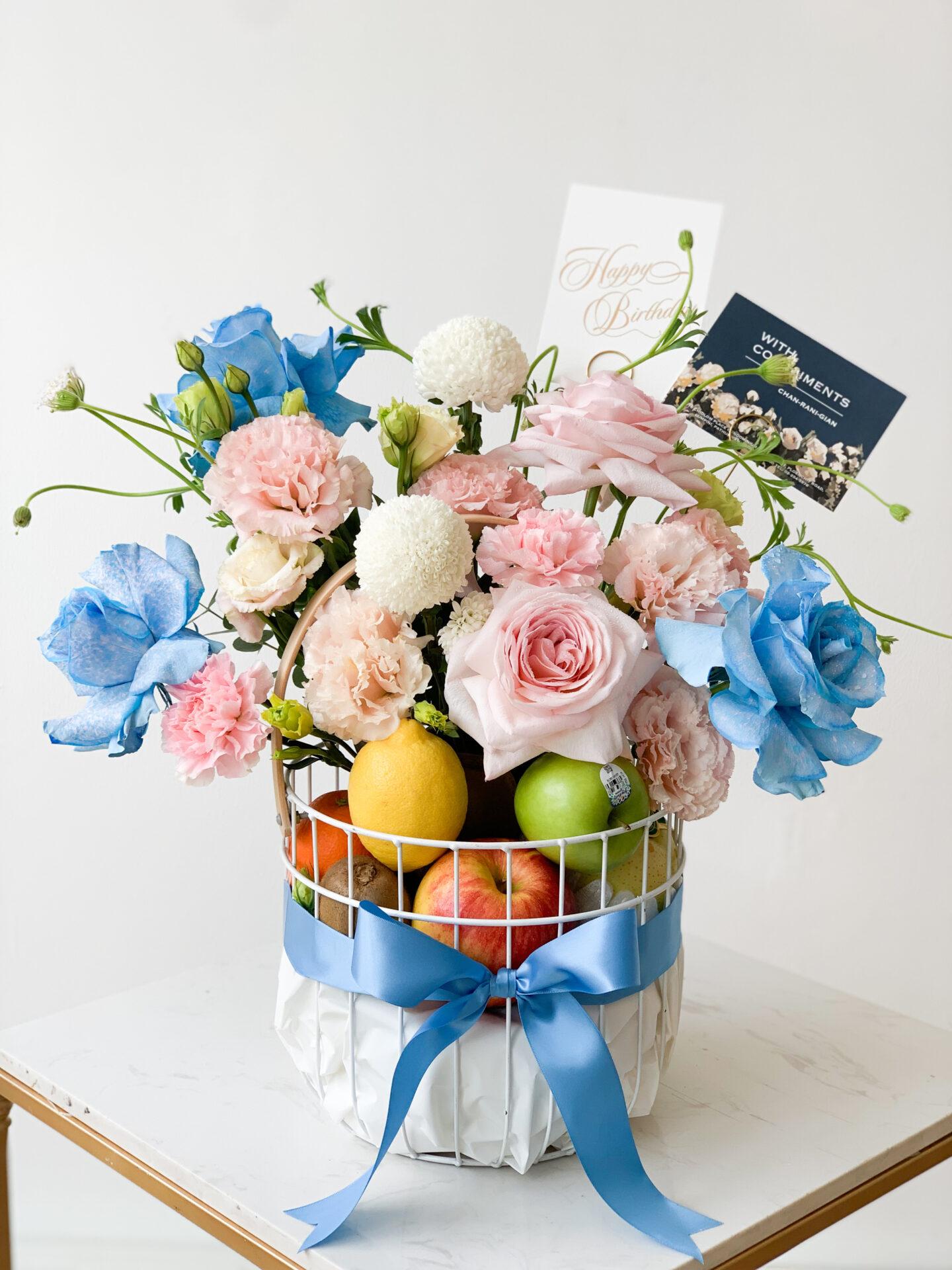 A flower arrangement in a white basket adorned with a blue ribbon. The bouquet features bright pink and blue flowers mixed with green and red apples. Two cards, one reading 