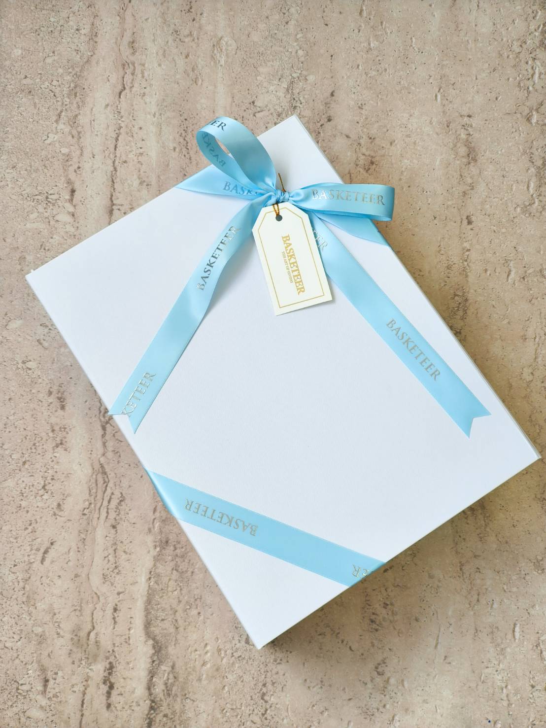 Elegant white gift box tied with pastel blue “Basketeer” ribbon and gift tag, luxury packaging for premium gifts.