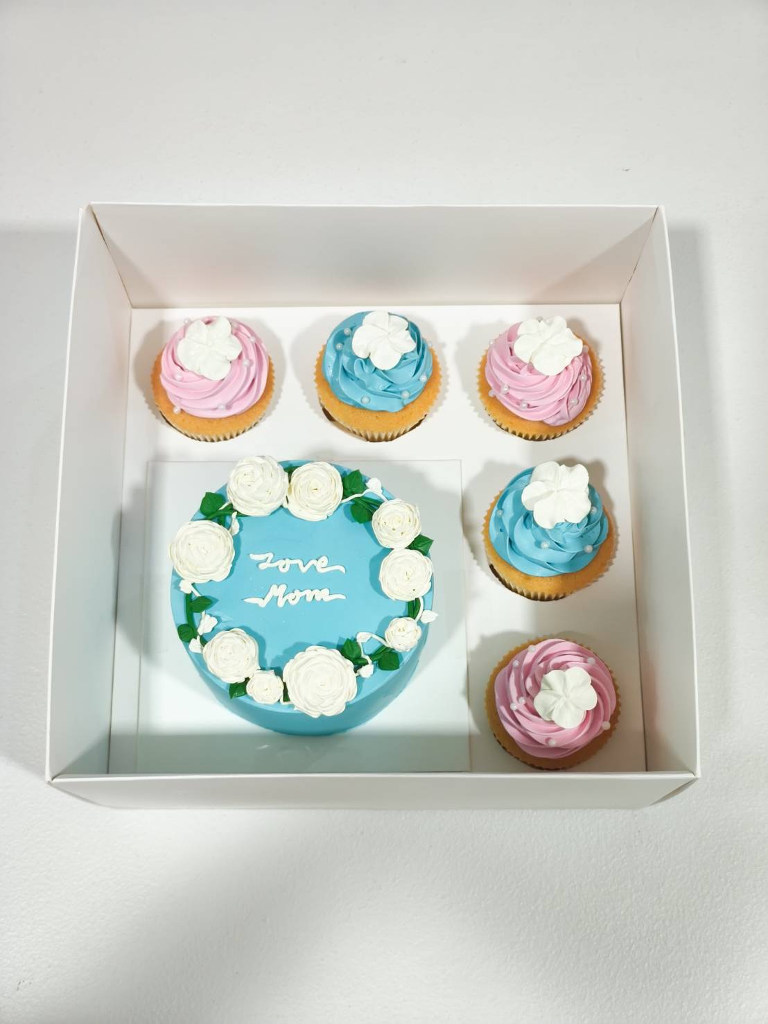 Mother’s Day gift set with blue frosted cake decorated with white roses and “Love Mom” message, paired with pink and blue frosted cupcakes in a gift box.