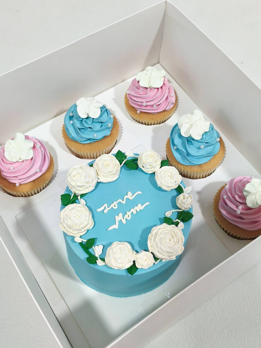 Angled view of Mother’s Day gift set with blue “Love Mom” cake decorated with white roses, paired with pink and blue frosted cupcakes in a gift box.