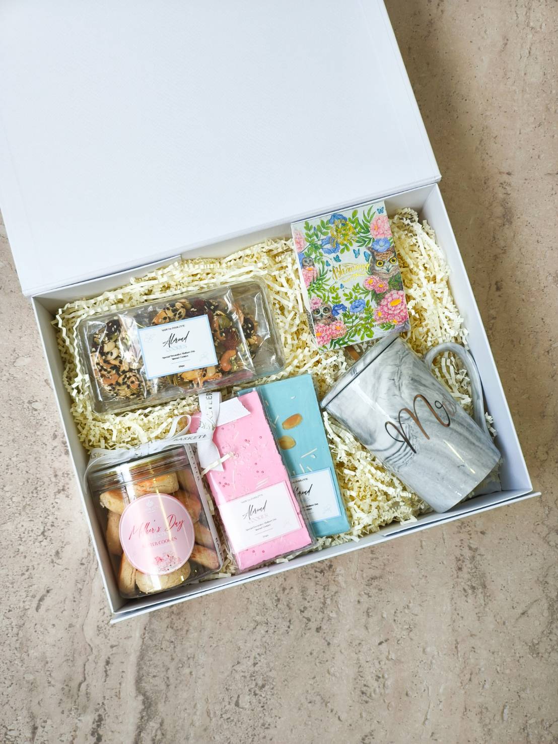 White gift box with assorted sweets, chocolate bars, cookies, a floral notebook, and a marble-patterned mug.