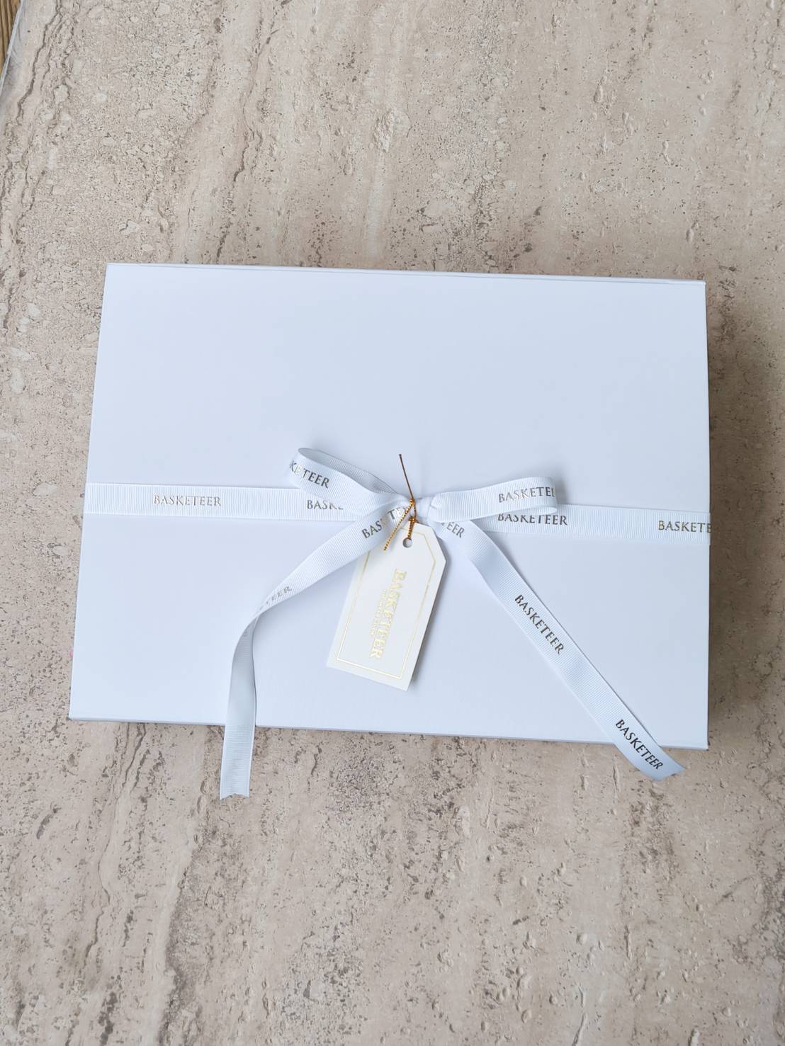 Elegant white magnetic gift box with white ribbon and gold-trimmed tag, placed on a marble surface.