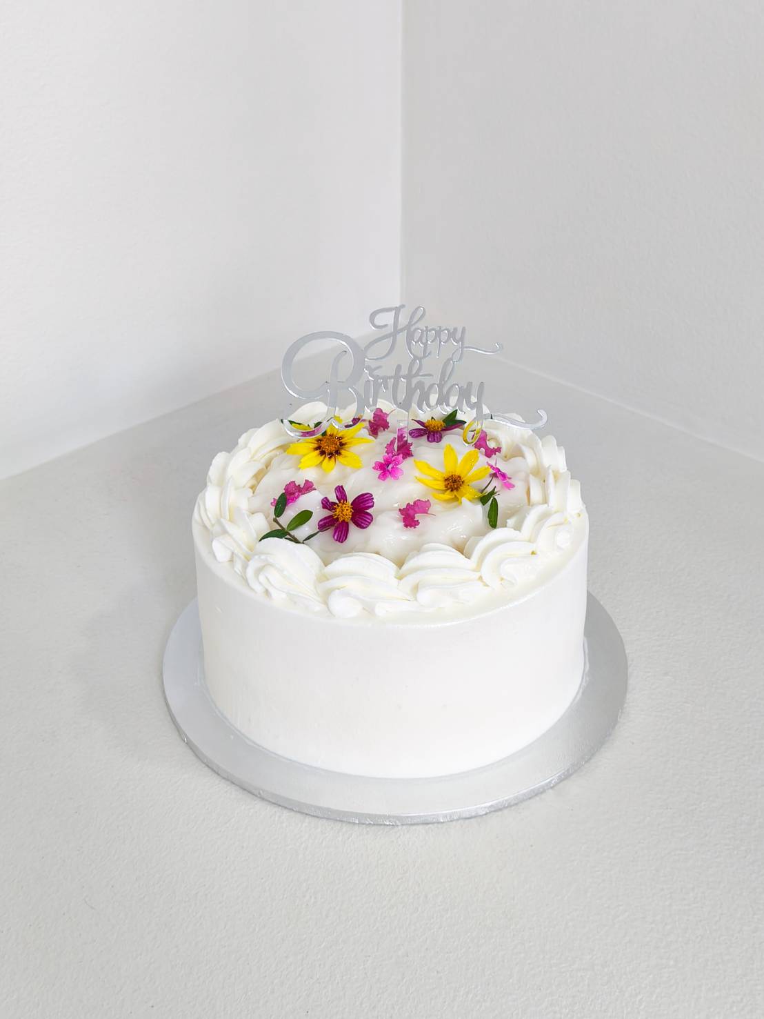 White cream cake decorated with fresh edible flowers in yellow and pink, topped with a silver Happy Birthday sign.