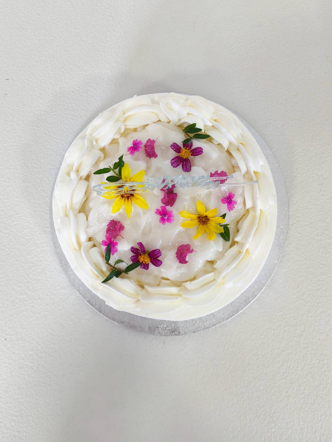 White cream cake top view decorated with fresh yellow and pink flowers, placed on a silver cake board.