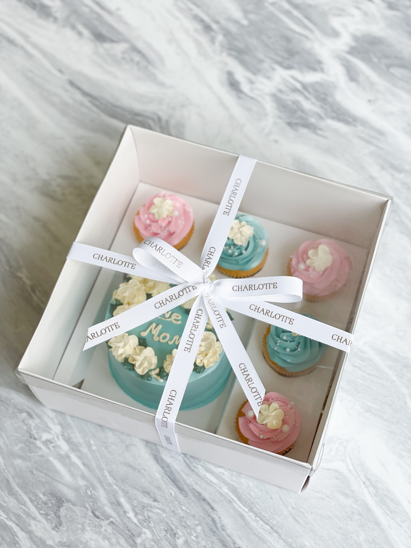 A square, pristine white box with a crystal-clear lid unveils an exquisite display: a delicately blue-frosted cake adorned with 