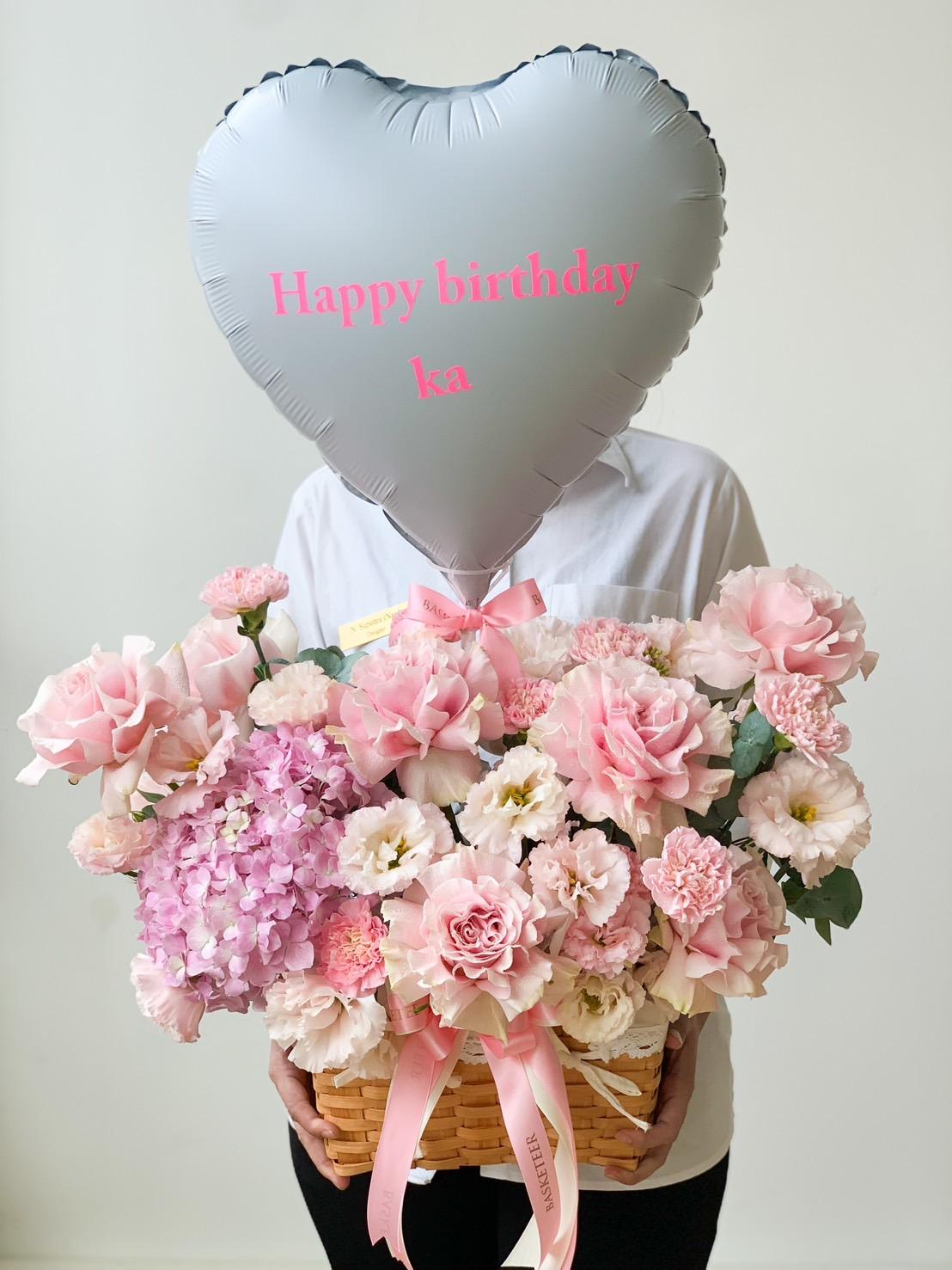 A person holding a basket filled with an assortment of pink and white flowers, showcasing Floral Elegance for Mother's Day. Attached to the basket is a metallic silver heart-shaped balloon with pink text that reads 