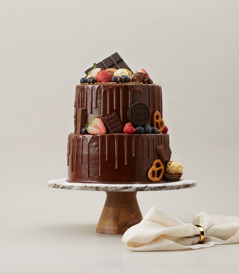 Two-tiered chocolate drip cake adorned with an assortment of toppings including fresh berries, chocolate bars, Oreo cookies, pretzels, and Ferrero Rocher chocolates, set on a marble cake stand.