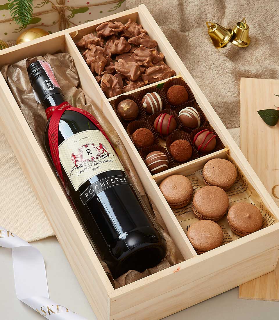 A refined wooden gift box by Basketeer showcases an exquisite bottle of red wine nestled amid a decadent assortment of chocolates, truffles, and caramel clusters. Savory macarons add an extra touch of sophistication to this luxurious ensemble. The box is opulently adorned with festive wrapping and elegant decorations, featuring a sleek ribbon displaying the Basketeer logo, charming bell ornaments, and a thoughtfully designed gift tag.