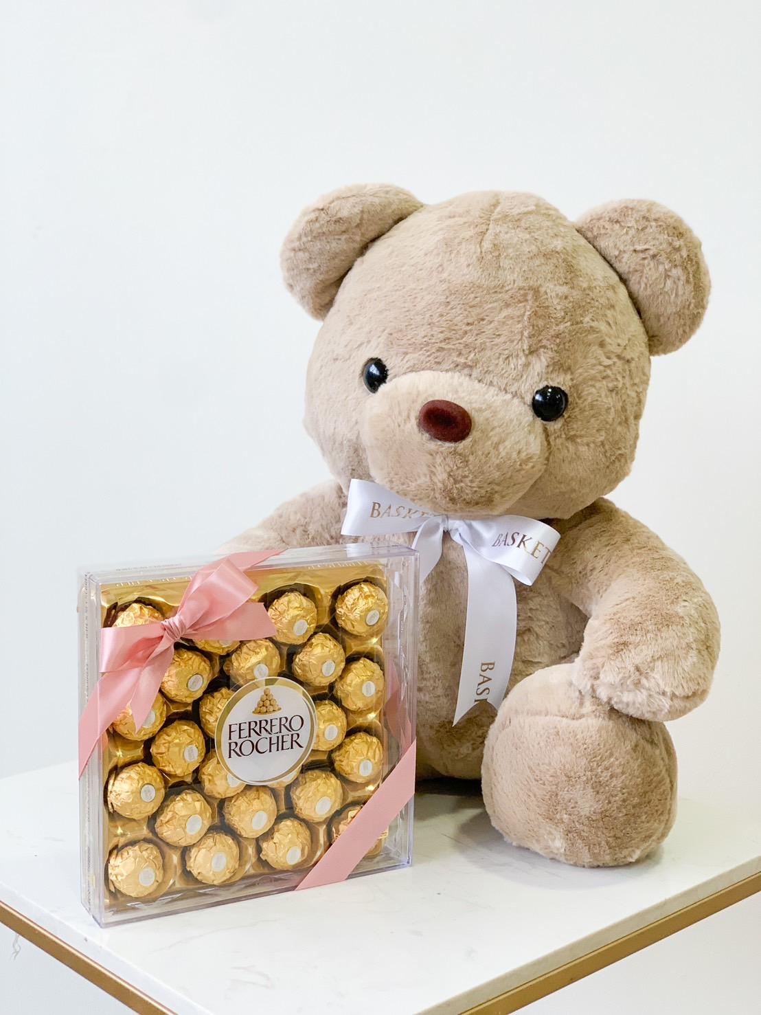 A large, fluffy teddy bear with a white bow sits on a surface next to a box of Ferrero Rocher chocolates. The chocolates, wrapped in gold foil and tied with a pink ribbon, make this sweet teddy chocolate gift absolutely delightful.