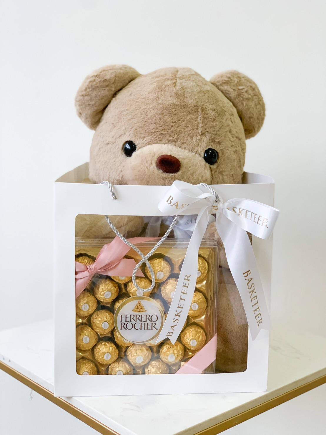 A large, fluffy teddy bear peeks out from a gift basket. The transparent section of the basket displays an enticing array of Ferrero Rocher chocolates. Adorned with white ribbons featuring 