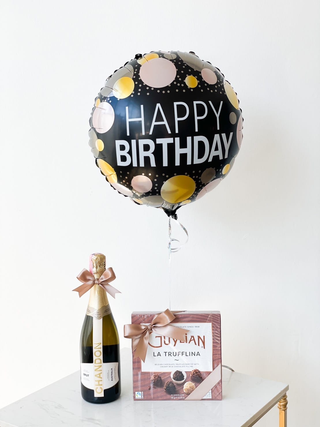 A black birthday balloon with "HAPPY BIRTHDAY" text, decorated with colorful dots, is tied to a bottle of Chandon sparkling wine and a box of Guylian La Trufflina chocolates. Both the bottle and the box have decorative bows, making it a perfect surprise gift combo set.
