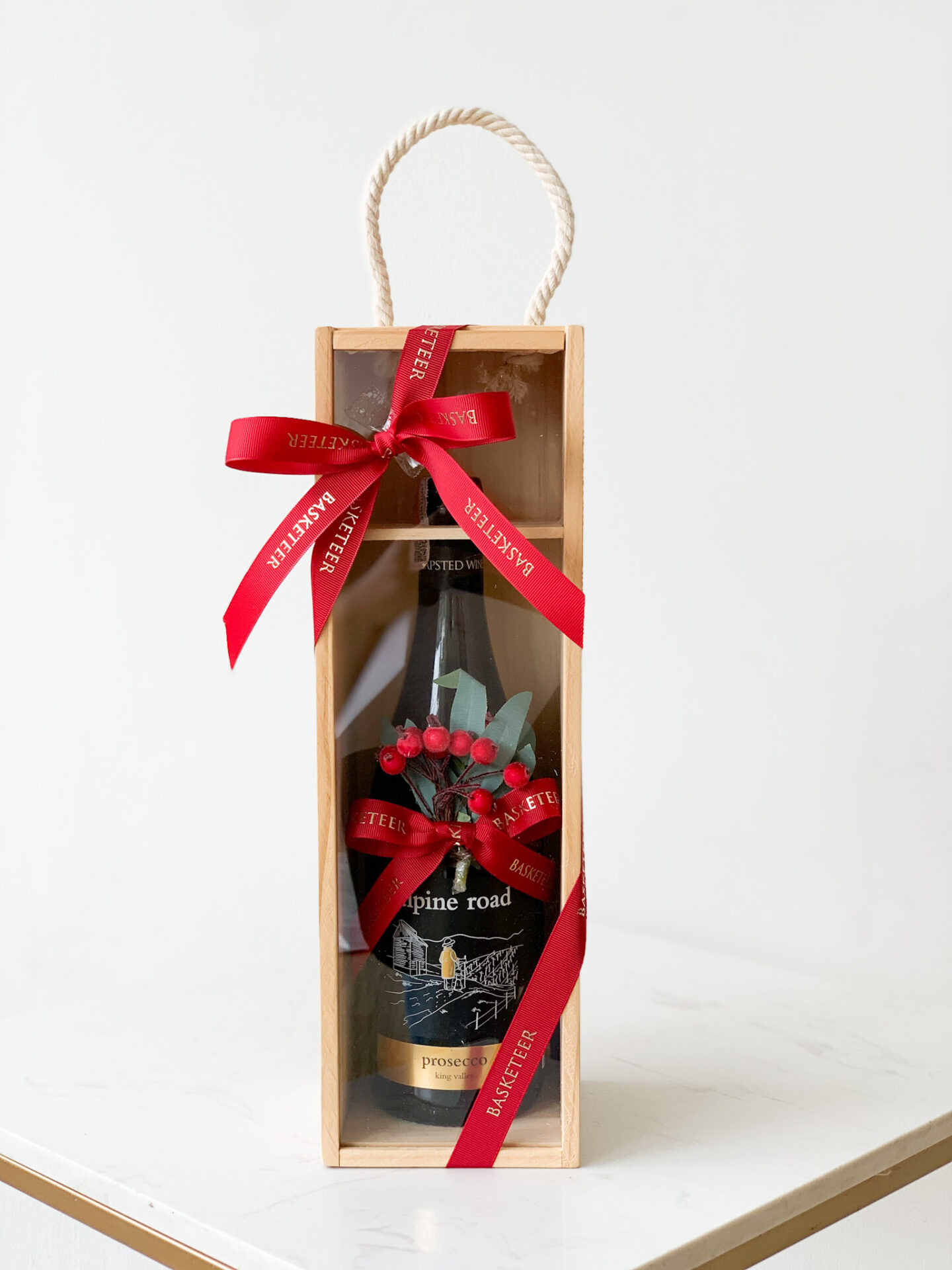 A bottle of "Pine Road Prosecco" in a wooden gift box with a rope handle. The box is adorned with a red ribbon and small decorative red berries and leaves, giving it a festive appearance perfect for gifting.