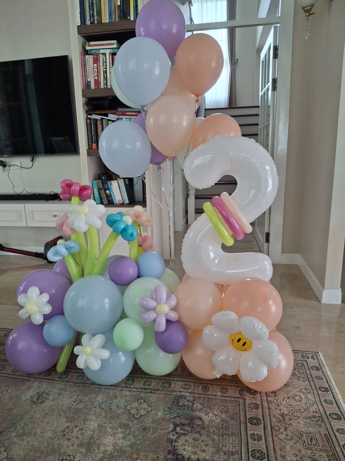 A pastel balloon display with a large white balloon shaped as the number 2, surrounded by flower-shaped balloons in pink, blue, purple, green, and a bee balloon on the floor in a home setting.