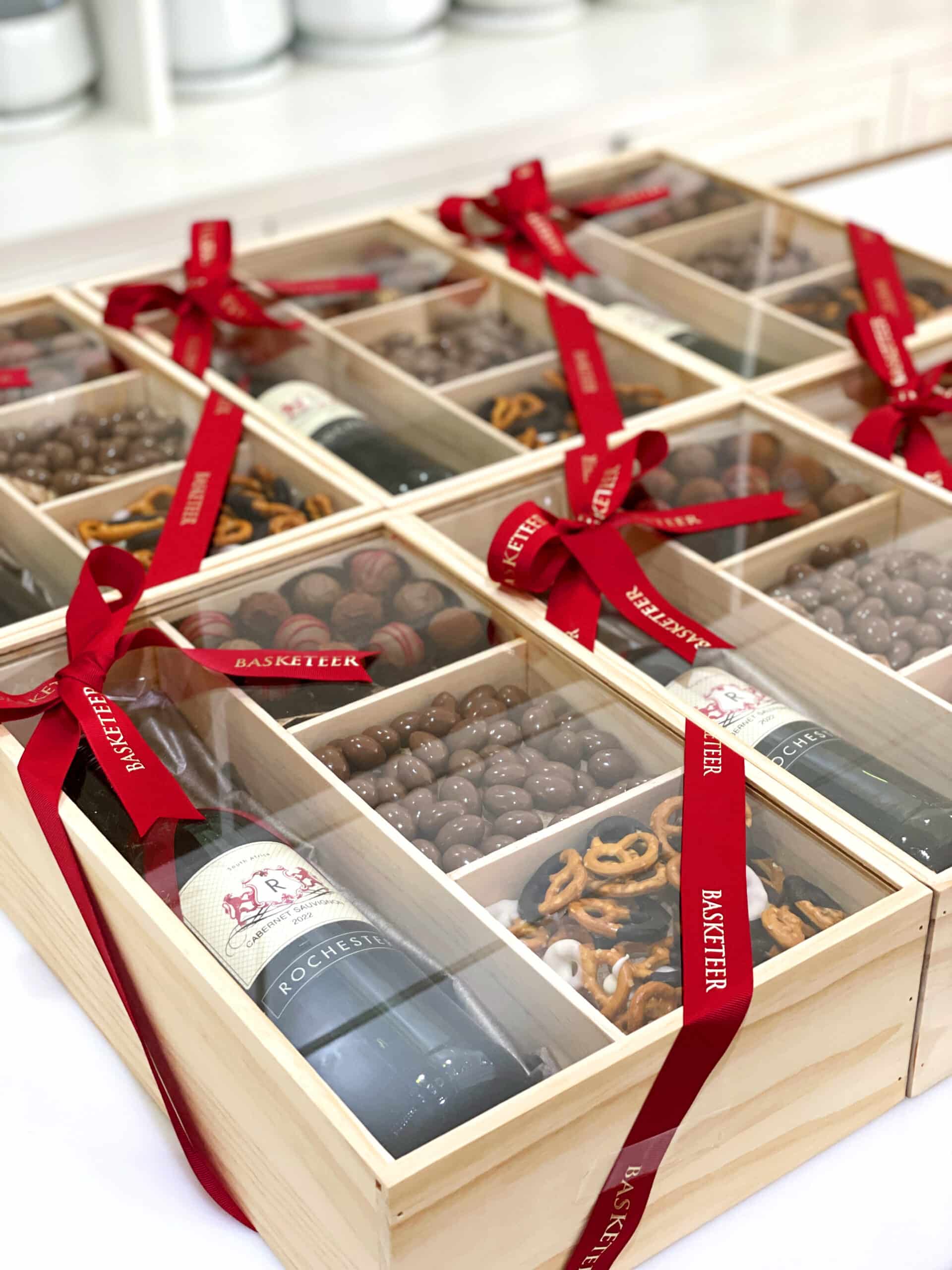 Wooden gift boxes containing a bottle of wine, assorted chocolates, and pretzels are arranged in a grid. Each box has a red ribbon with "Basketeer" written on it, tied into a bow.