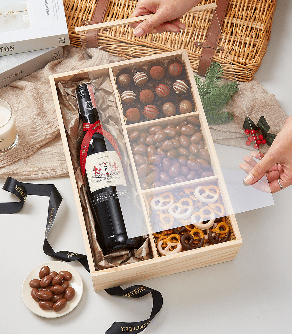 A person is opening a wooden gift box containing a bottle of wine, assorted chocolate truffles, chocolate-covered nuts, and chocolate-covered pretzels. There are festive decorations, a candle, a book, and a ribbon on the table, along with a wicker basket.