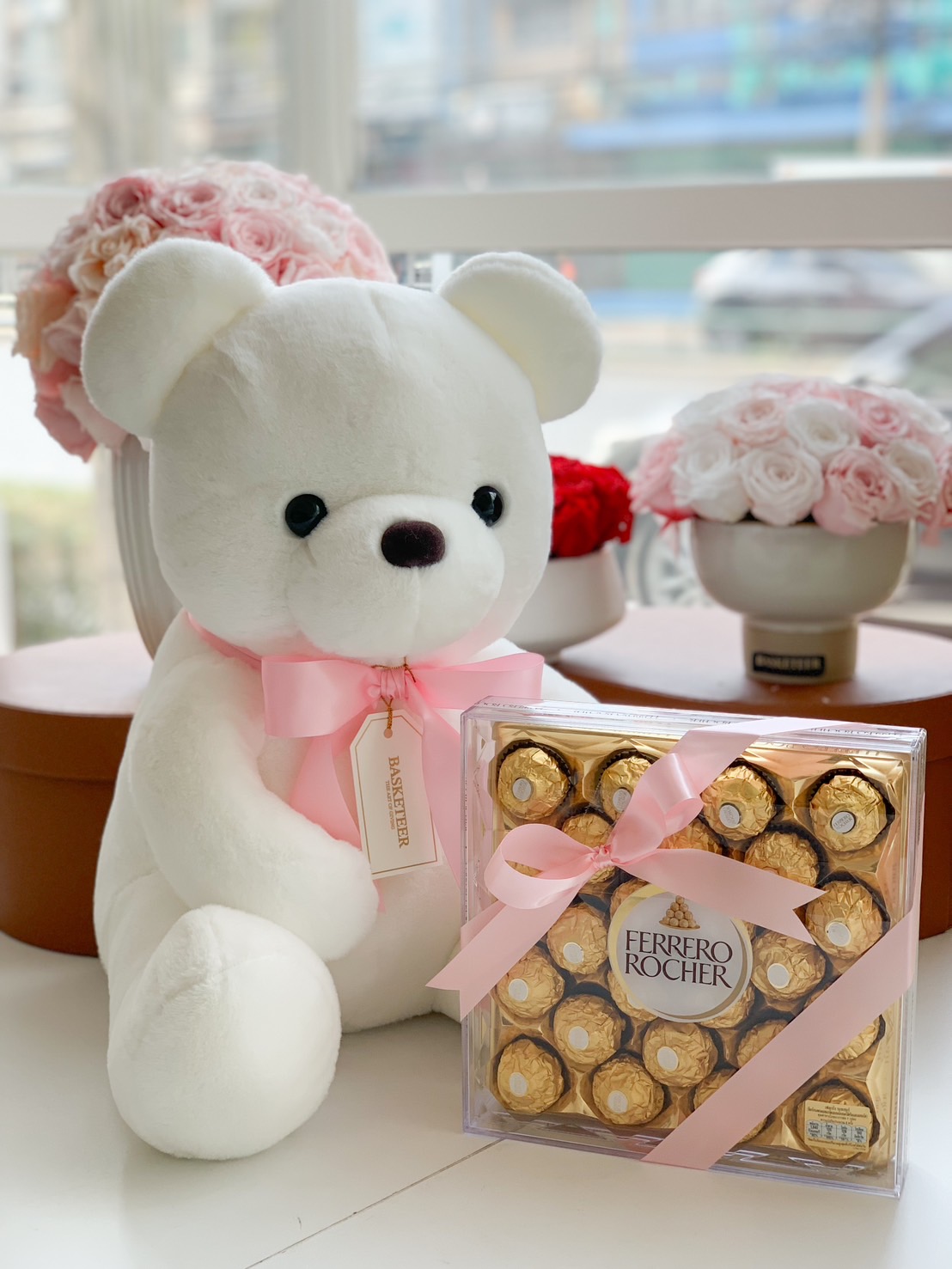 A plush white teddy bear with a pink ribbon and a luxurious box of Ferrero Rocher chocolates, elegantly wrapped with a pink satin bow, creating the perfect romantic or heartfelt gift.