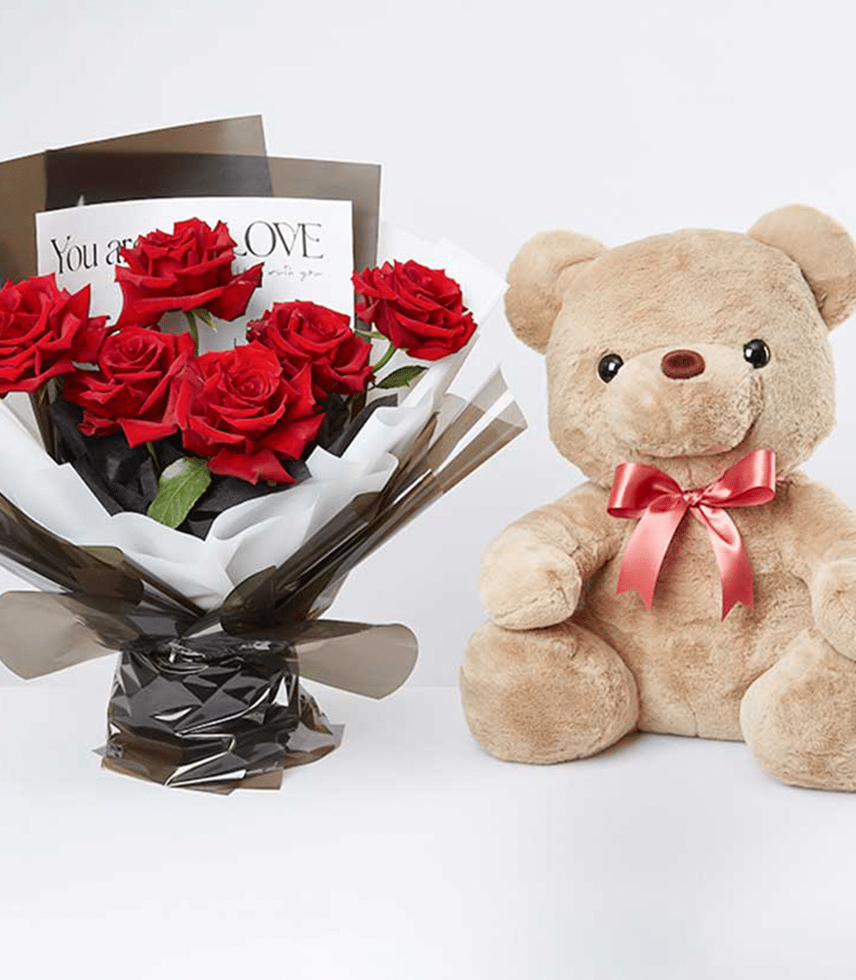 A lavish bouquet of velvety, crimson roses elegantly ensconced in luxurious dark and white wrapping paper graces the scene. Beside this opulent floral arrangement rests an oversized, plush beige teddy bear, adorned with a delicate pink satin ribbon around its neck. The sophisticated ensemble includes a refined card featuring heartfelt text, epitomizing thoughtful luxury. Presented against a pristine white background, these exquisite offerings are from none other than Basketeer, where sophistication meets sentimentality.