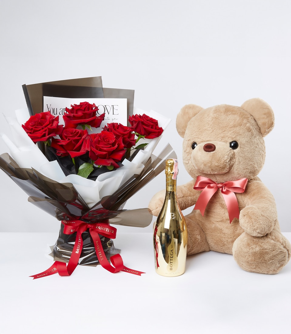 A luxurious bouquet of exquisite red roses, elegantly wrapped in white and black paper adorned with a sumptuous red ribbon that proudly displays the Basketeer logo. Nestled beside is a charming, large beige teddy bear, its neck graced with a rich red ribbon. Completing this opulent arrangement is a bottle of premium gold-colored champagne, all meticulously presented on an immaculate white surface—a scene set for pure indulgence and delight courtesy of Basketeer.