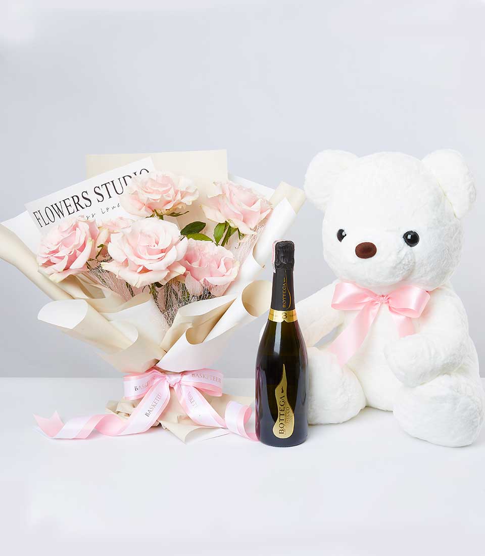 A sumptuous bouquet of pink roses ensconced in elegant beige paper, accompanied by an exquisite bottle of sparkling wine and a grand white teddy bear adorned with a delicate pink ribbon around its neck. The bouquet features a refined 