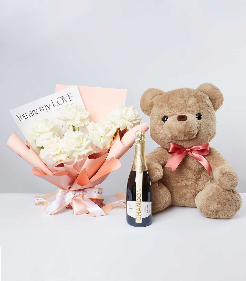 A sumptuous plush teddy bear adorned with a delicate pink bow nestles beside an elegant bouquet of pristine white roses, luxuriously wrapped in peach-hued paper. Accompanying this splendid arrangement is a card with the heartfelt message 