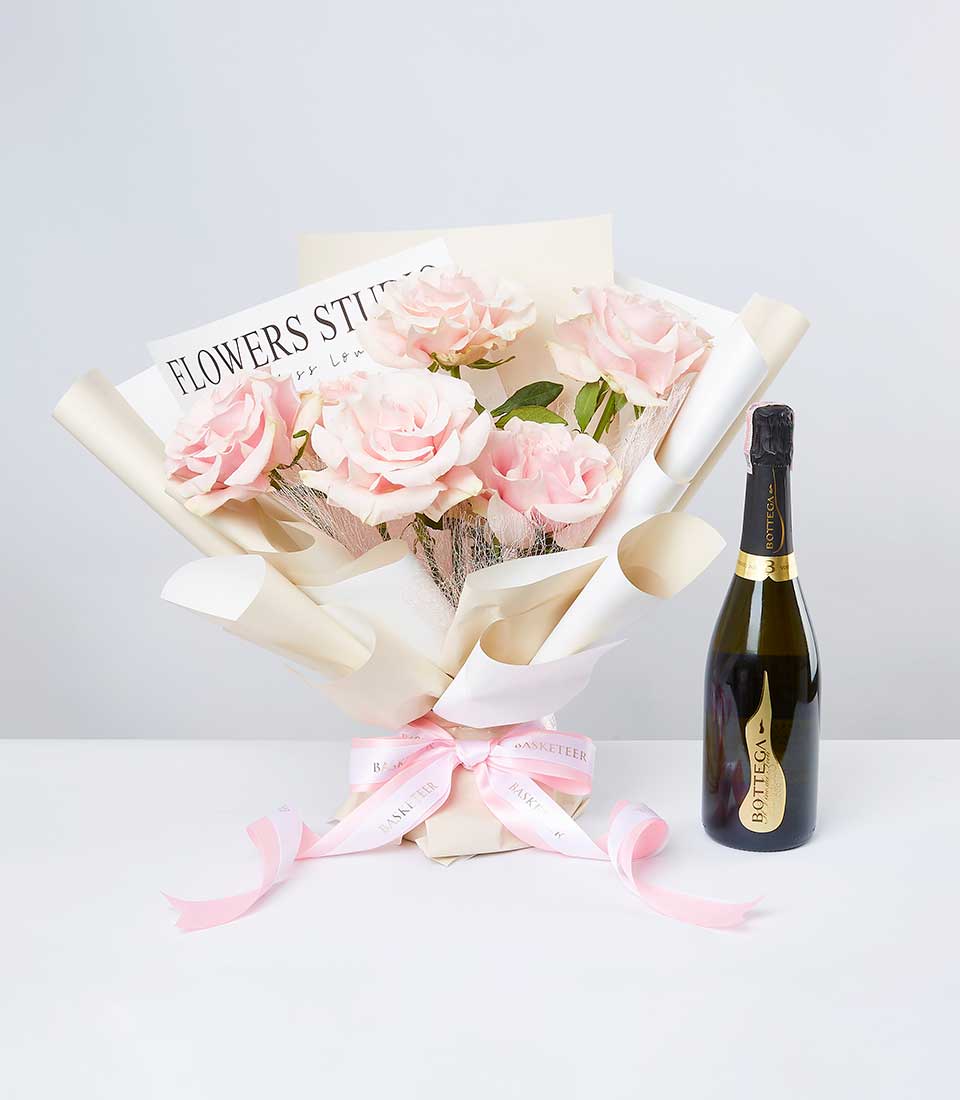 A lavish bouquet of blush pink roses, gracefully enveloped in elegant cream-colored paper and adorned with a sophisticated pink ribbon bearing the Basketeer logo, is tastefully arranged next to a chic bottle of Bottega sparkling wine. A stylish card from 