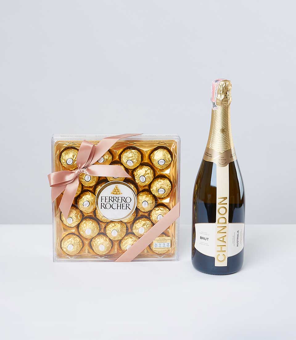 A luxurious Basketeer gift box adorned with a delicate pink ribbon and bow, showcasing an exquisite assortment of Ferrero Rocher chocolates. Nestled beside this gourmet treat is a refined bottle of Chandon Brut sparkling wine, elegantly wrapped in golden foil and bearing a pristine white label with the prestigious 