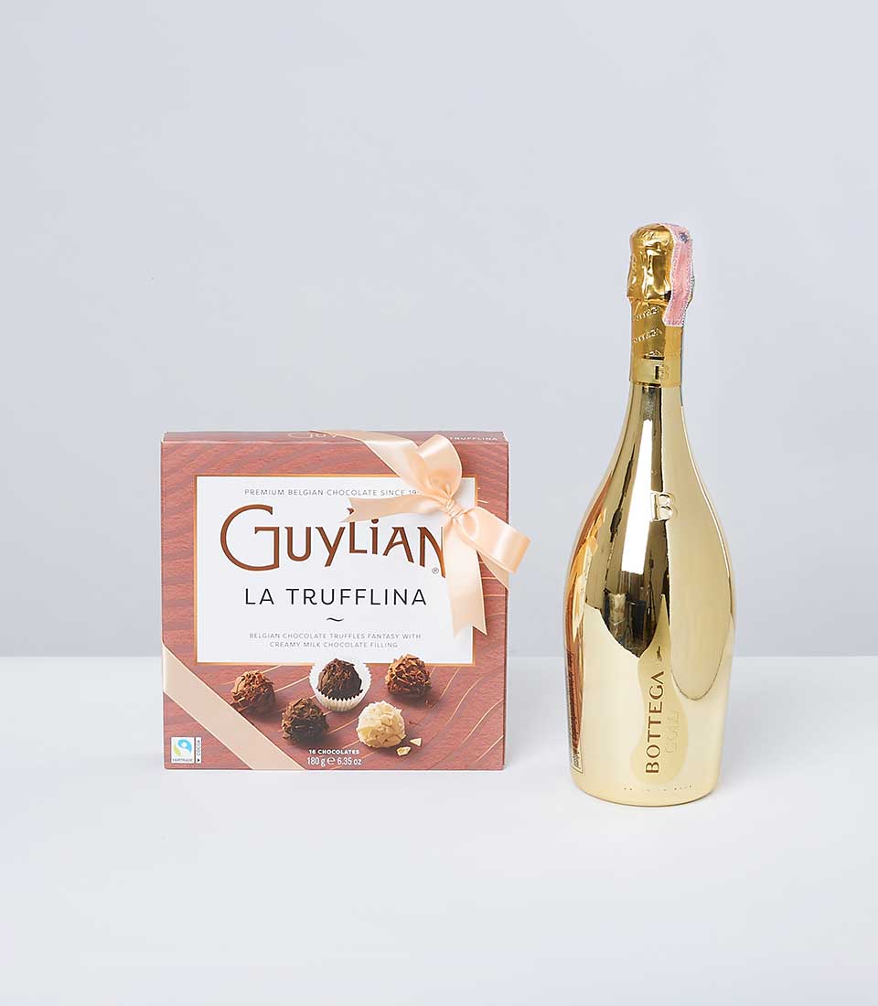 A luxurious gold bottle of Bottega sparkling wine stands elegantly beside a sophisticated rectangular box of Guylian La Trufflina chocolates. The box showcases an exquisite image of assorted truffles and is beautifully adorned with a peach-colored ribbon tied in a graceful bow at the top left corner. The scene is completed with the touch of opulence from Basketeer, suggesting indulgence and refined taste.