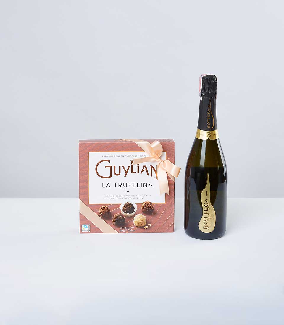 A luxurious presentation featuring a box of Guylian La Trufflina chocolates, elegantly wrapped in a light pink ribbon bearing the Basketeer logo. Beside it, a glistening bottle of Bottega Gold sparkling wine sits regally on a pristine white surface, all set against an understated light gray backdrop. The scene exudes refined elegance and sophistication, perfect for indulging in pure luxury.
