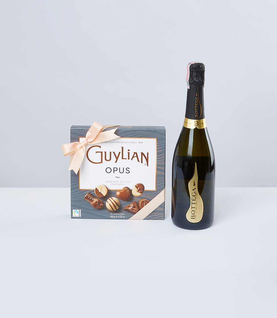 A tastefully arranged box of Guylian Opus assorted Belgian chocolates, adorned with a delicate peach-colored ribbon featuring the Basketeer logo, stands elegantly beside a bottle of Bottega Prosecco. The luxurious ensemble is set against a sophisticated light gray backdrop, inviting indulgence and refinement.
