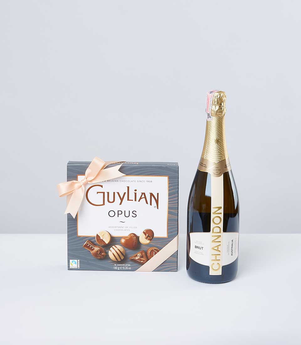 A meticulously crafted box of Guylian Opus assorted chocolates, elegantly adorned with a delicate light pink ribbon bearing the prestigious Basketeer logo, is positioned beside a sumptuous bottle of Chandon Brut sparkling wine. These exquisite delights are tastefully arranged against a pristine white backdrop, capturing an air of refined luxury and inviting indulgence.
