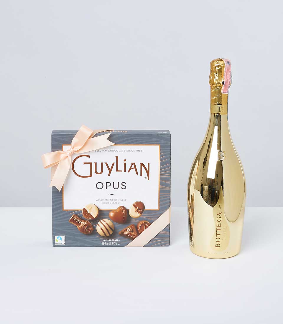 A luxurious bottle of Bottega Gold Prosecco gleams alluringly beside an exquisite box of Guylian Opus assorted Belgian chocolates. The chocolate box, adorned with a sumptuous peach-colored ribbon bow, showcases the prestigious Basketeer logo, promising an indulgent experience. The elegant arrangement is set against a sophisticated light gray backdrop, inviting you to indulge in pure elegance and taste.