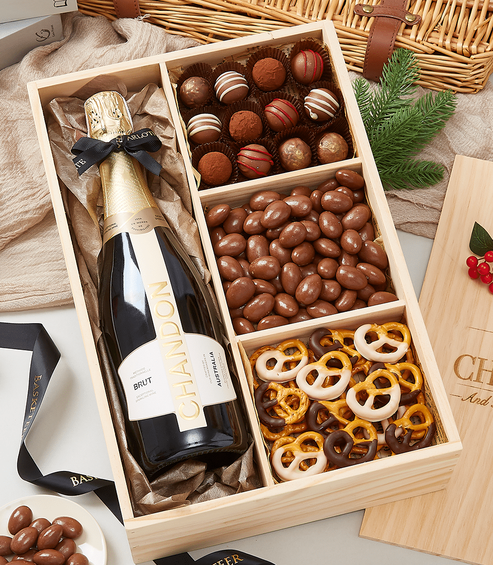 A luxurious wooden gift box by Basketeer holding an exquisite bottle of Chandon Brut champagne, elegantly nestled amidst a selection of decadent chocolates. Delicate truffles, luscious chocolate-covered almonds, and gourmet chocolate-drizzled pretzels enhance the opulent array. This inviting display is set on a rustic surface adorned with a festive green pine sprig, promising an indulgent experience. The Basketeer logo graces the ribbon and box, underscoring the superior quality of this lavish gift.