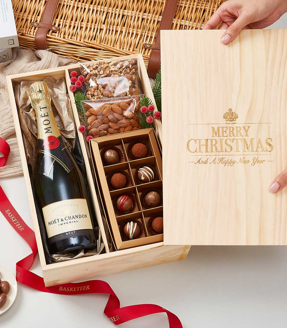 A tastefully designed wooden gift box partially open, unveiling an opulent bottle of Moët & Chandon champagne, decadent chocolate truffles, a deluxe container of mixed nuts, and elegant festive decorations. The lid of the box is meticulously engraved with 