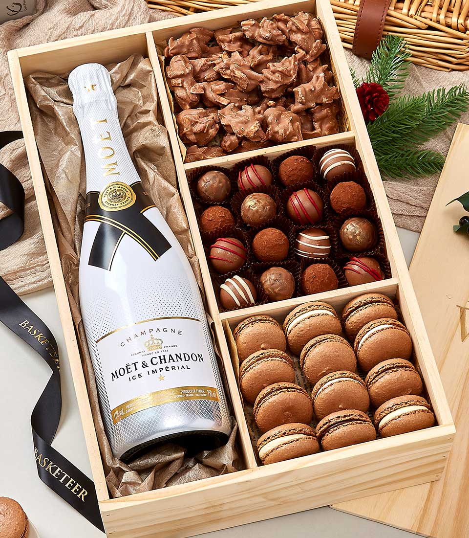 A luxurious wooden gift box elegantly displays a bottle of Moët & Chandon Ice Imperial champagne, surrounded by an assortment of premium chocolates and exquisitely arranged macarons. The interior is thoughtfully adorned with decorative paper shavings, enhancing the presentation. The entire ensemble is accented by a sophisticated ribbon featuring the distinguished Basketeer logo, epitomizing indulgence and exclusivity.