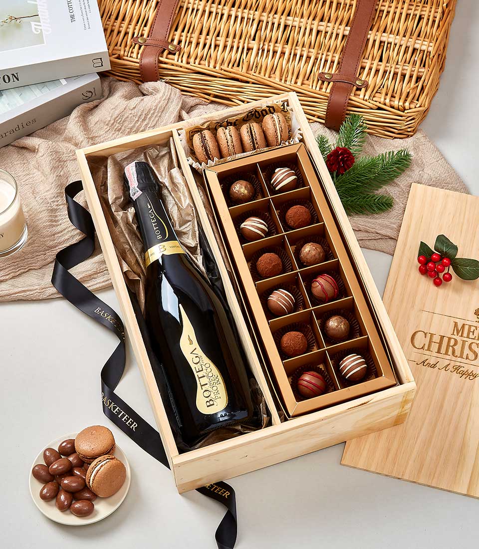 A luxurious wooden gift box from Basketeer, elegantly adorned with a black ribbon bearing the Basketeer logo. Inside, you'll find a fine bottle of sparkling wine nestled among an exquisite selection of assorted chocolates and vibrant macarons. In the background, a charming wicker basket adds rustic sophistication to the scene. Nearby, additional macarons await, accompanied by a flickering candle that casts a warm glow and a Merry Christmas sign embellished with festive holly – creating an inviting holiday atmosphere.