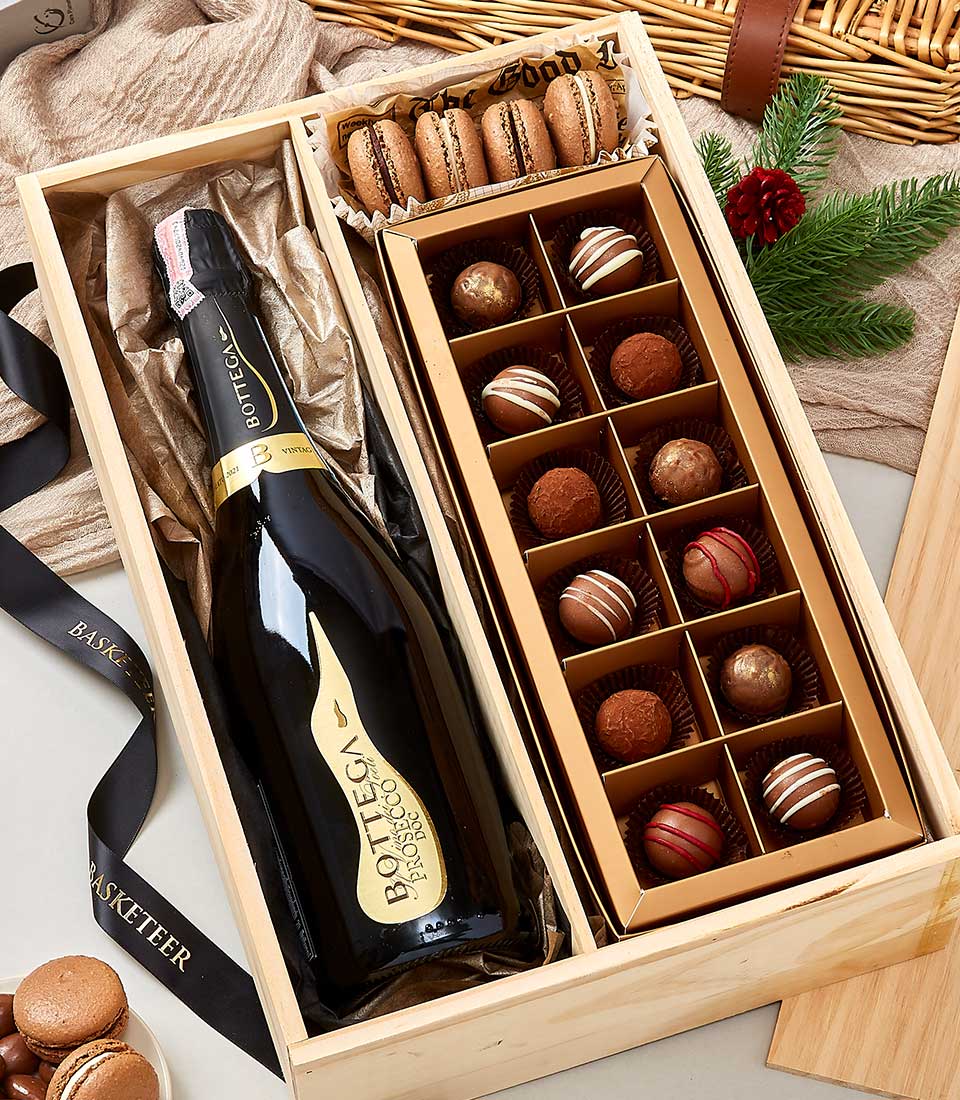 A luxurious wooden gift box from Basketeer, featuring an elegant bottle of Bottega Prosecco, a colorful array of assorted macarons, and a meticulously arranged selection of 15 gourmet chocolate truffles. The presentation is enhanced with festive touches including a sprig of pine and delicate pinecones. In the background, a charming wicker basket and Basketeer-branded ribbon add to the opulent ambiance.