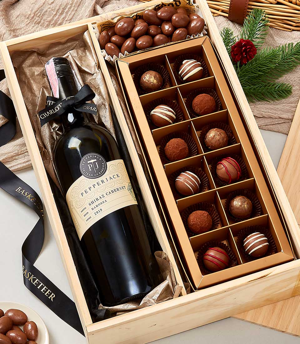 A lavish wooden gift box from Basketeer featuring an exquisite bottle of Pepperjack Shiraz Cabernet wine elegantly positioned on the left and a curated selection of decadent assorted chocolates on the right. The scene is beautifully embellished with a festive ribbon adorned with the Basketeer logo, and accented by a sprig of pine, creating an inviting holiday atmosphere.
