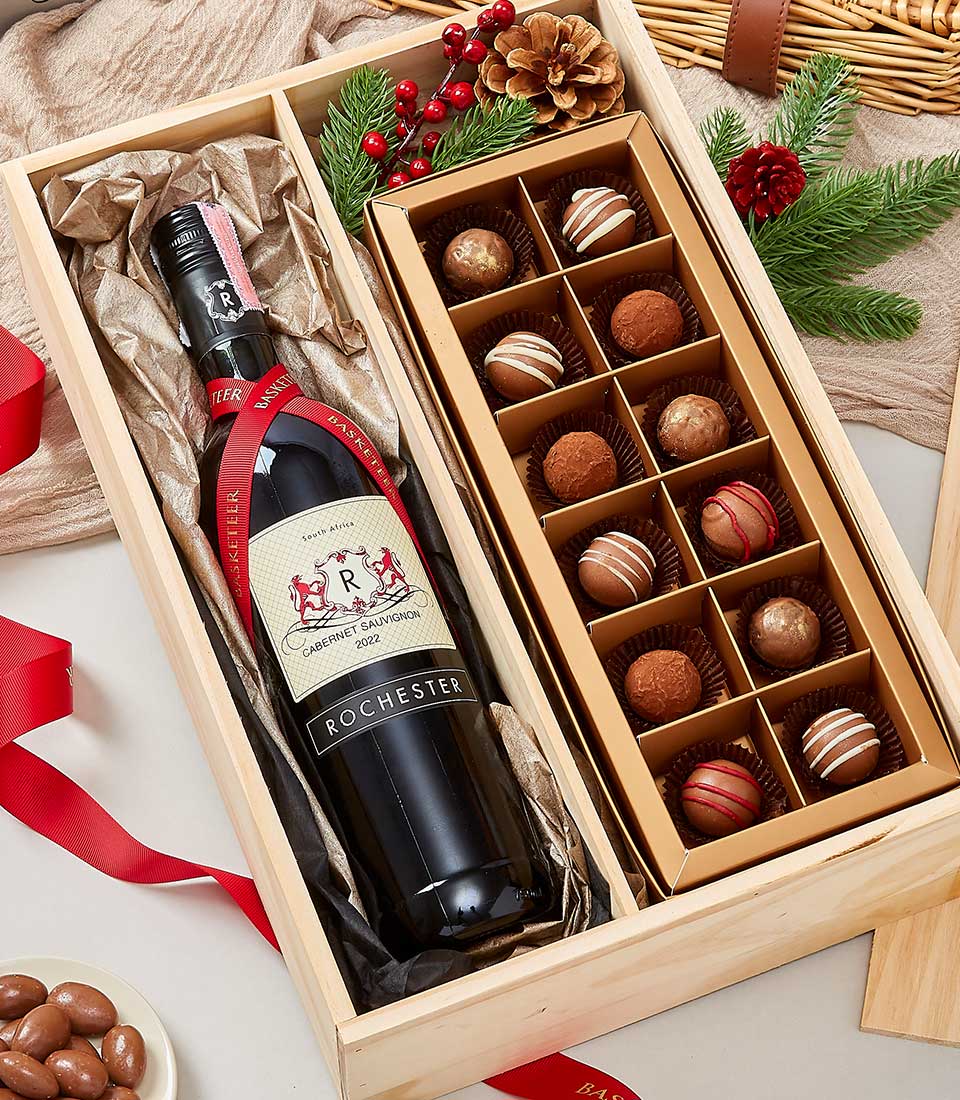 A luxurious Basketeer gift box elegantly adorned with pine cones, lush fir branches, and a sophisticated red ribbon. Inside, discover the exquisite Rochester Cabernet Sauvignon 2022 paired with an indulgent selection of gourmet chocolates. This impeccable presentation and sumptuous contents make it the ultimate holiday gift set.