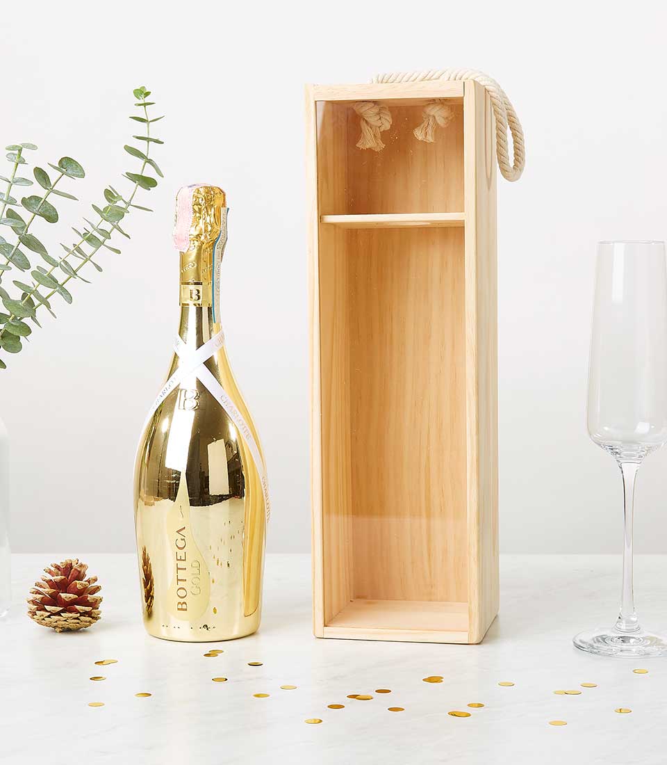 A luxurious gold bottle of Bottega Prosecco Gold Box stands elegantly beside an empty wooden wine box adorned with a rope handle, exuding charm and sophistication. A crystal-clear champagne flute sparkles to the right, while delicate pine cones and shimmering gold confetti sprinkle the pristine white surface, creating a scene of festive opulence. Graceful eucalyptus stems add a natural touch in the background, enhancing the inviting atmosphere. The entire setting whispers luxury and refinement, perfect for any special occasion hosted by Basketeer.