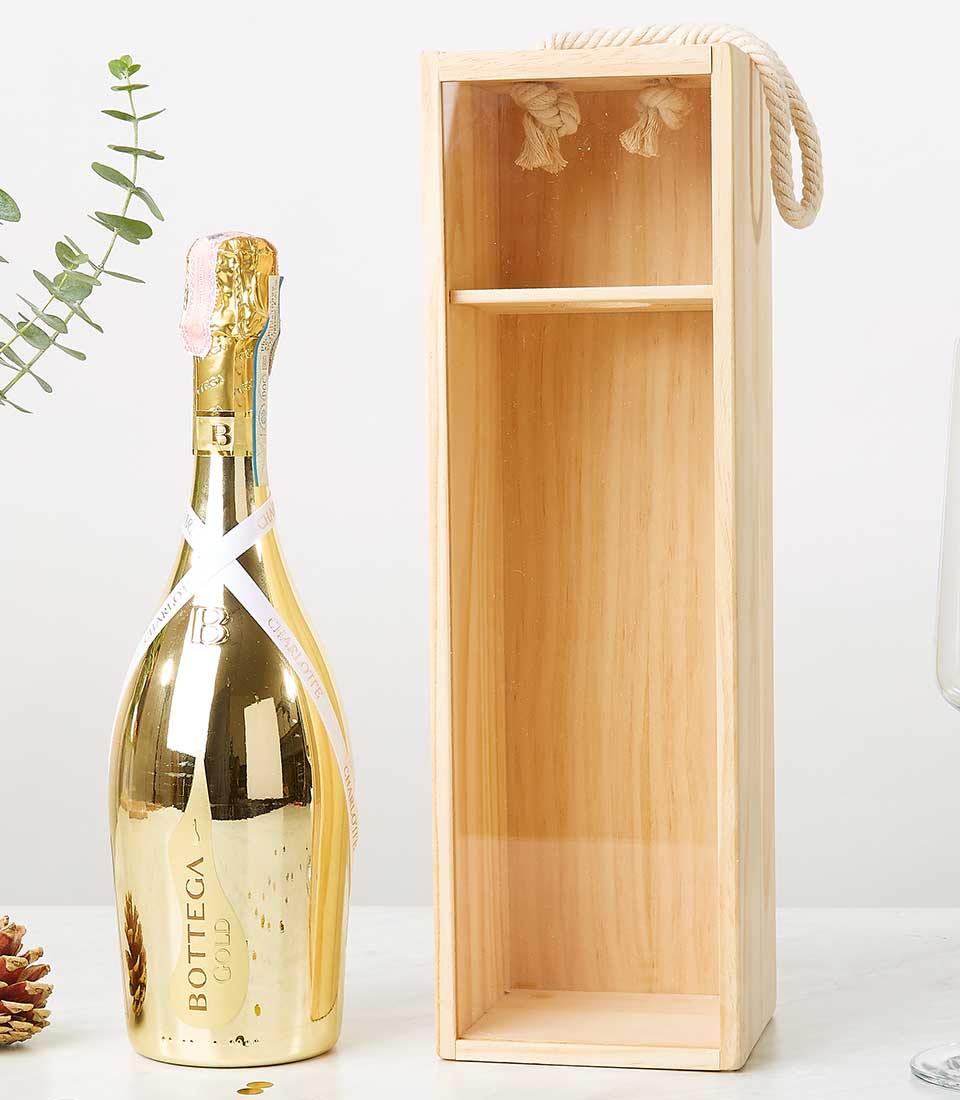 A gold Bottega Prosecco bottle stands proudly beside an opulent, empty Bottega Prosecco Gold Box adorned with an elegant rope handle, showcasing the height of sophistication. The setting is complemented by a crystal glass and a touch of nature featuring lush green leaves and a glistening pine cone, crafting a festive and luxurious ambience. A ribbon graced with the Basketeer logo adds an extra layer of premium charm to this exquisite scene.
