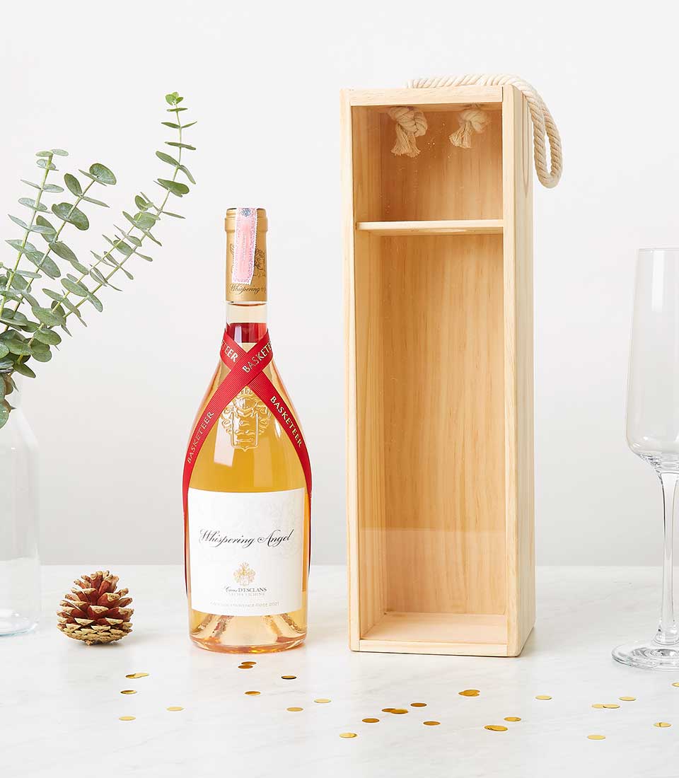 A bottle of Whispering Angel rosé wine exudes sophistication as it stands elegantly beside a luxe wooden gift box, adorned with the distinguished Basketeer logo and a chic rope handle. The scene beckons with an inviting empty wine glass, a charming pine cone, and a delicate sprig of eucalyptus. Adding to the festive ambiance, scattered gold confetti sparkles on the pristine white surface, creating an atmosphere of refined celebration.