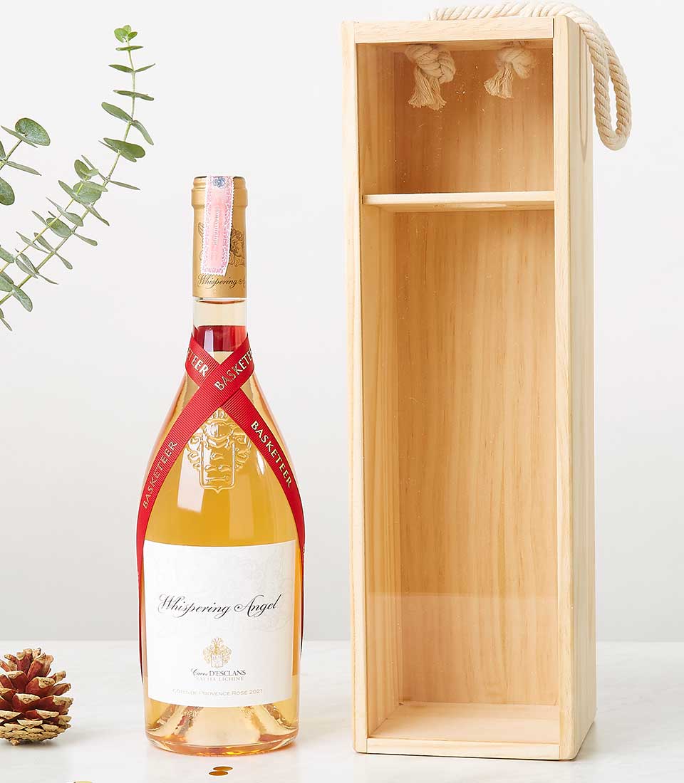 A luxurious presentation featuring a bottle of Whispering Angel rosé wine elegantly tied with a rich red ribbon. The exquisite bottle rests next to an open wooden gift box, complete with the distinguished Basketeer logo. To enhance its allure, delicate green leaves and an artfully placed pinecone add a touch of nature's charm. The gift box, designed for opulence, boasts a sturdy rope handle, ready for effortless gifting or indulgence.
