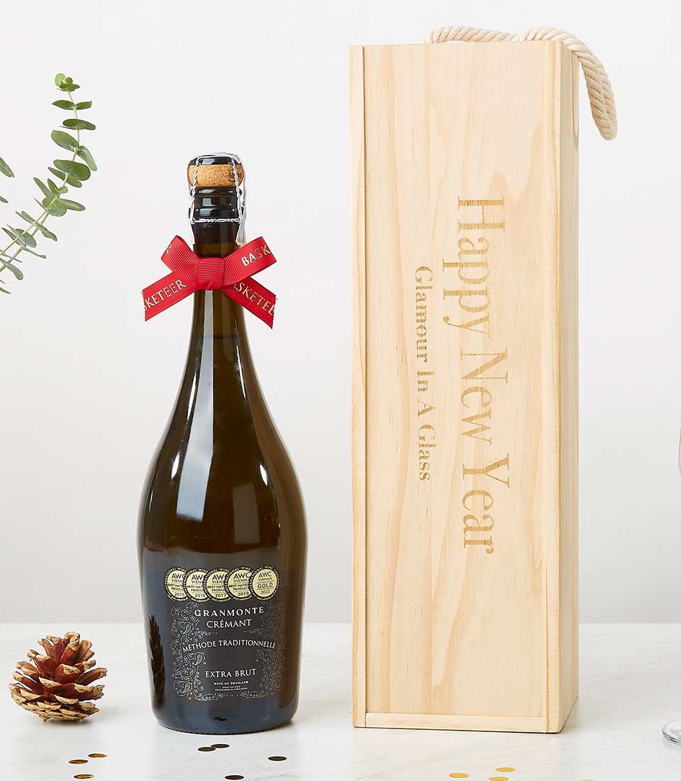 A bottle of Gramont Crémant elegantly adorned with a red ribbon around the neck stands beside a luxurious wooden gift box, engraved with the words 